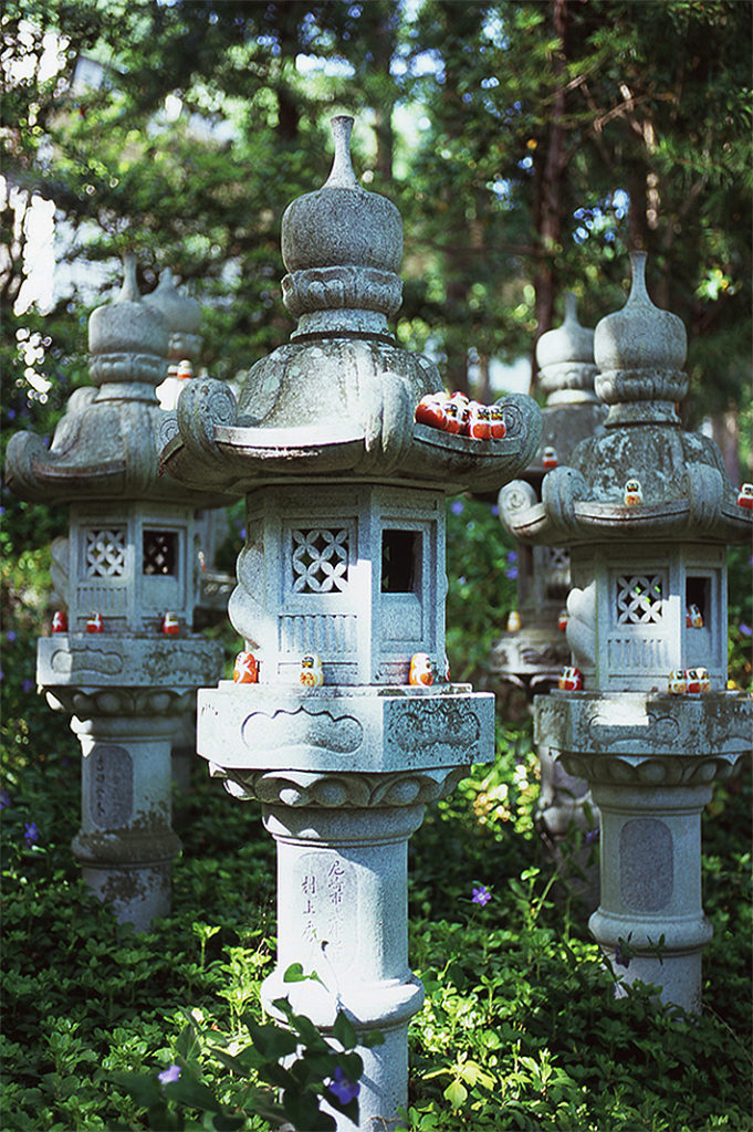 Tiny wooden Daruma sit dotted across three stone lanterns, backed in the green woodland of Katsuō-ji.