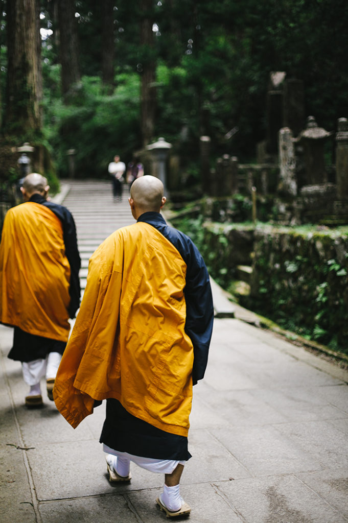 Buddhist monks walk away from the mausoleum as two Japanese pilgrims stop to observe, deep in the cedar forests of Kōya-san.