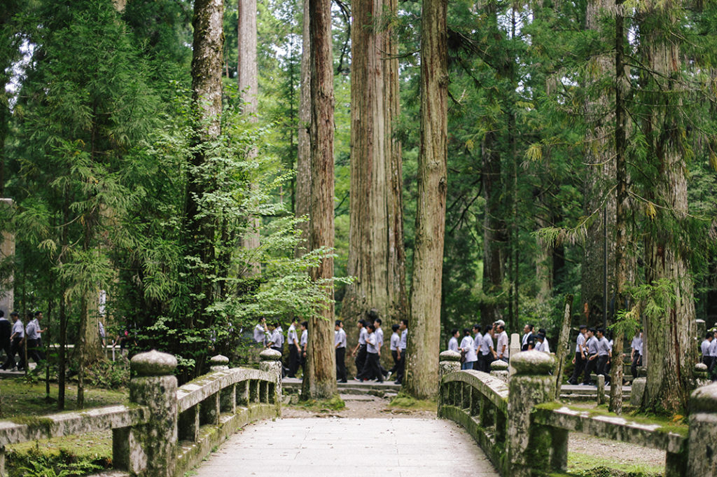 A sea of white-shirted schoolchildren walk in a line along a pathway shrouded in trees, set behind a stone bridge deep in Kōya-san's Okuno-in.