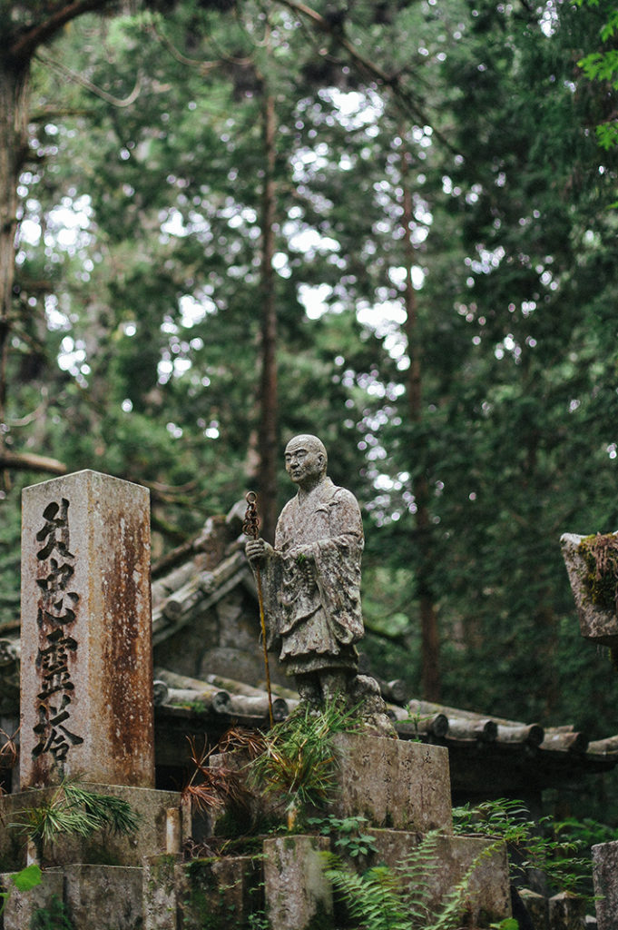 A stone statue of the Buddhist master Kukai stands surrounded by cedar trees and ferns in Kōya-san.